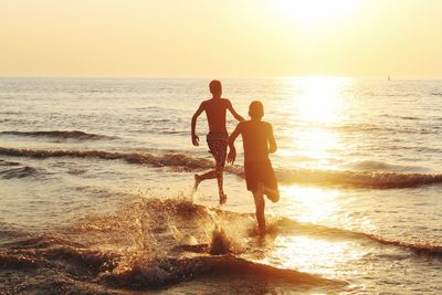 Rear view of silhouette friends running in sea against sky during sunset