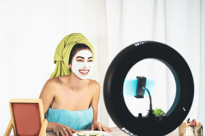 Smiling young woman with facial mask filming over smart phone
