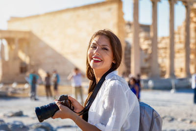 Portrait of happy young woman photographing with camera against old ruins
