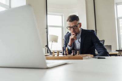Businessman looking at laptop while playing chess at office
