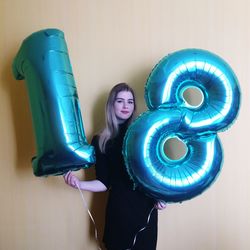 Portrait of young woman holding number helium balloons against wall