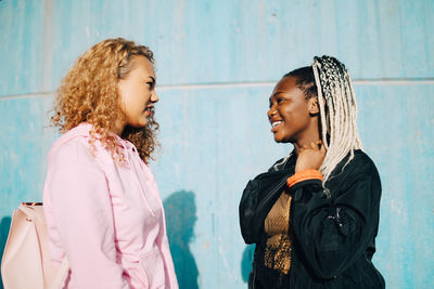 Smiling female friends talking while standing by wall during sunny day