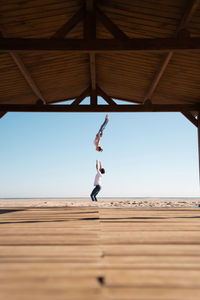 Side view of distant unrecognizable man throwing female acrobat up in air performing somersault on sandy shore on summer day