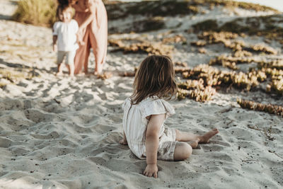 Behind view of young toddler girl sitting in sand looking at family