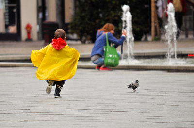 Full length rear view of boy running behind pigeon on street in city