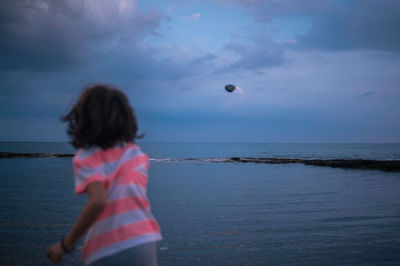Rear view of girl throwing stone towards sea against cloudy sky