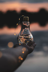 Cropped hand holding illuminated string lights in bulb at sunset