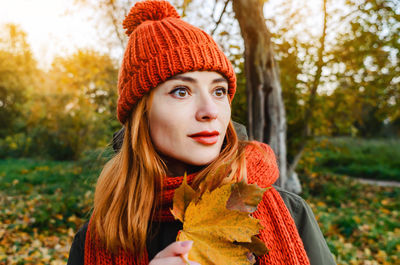 Portrait of mysterious woman with autumn yellow leaf in hand in orange knitted hat and scarf. 
