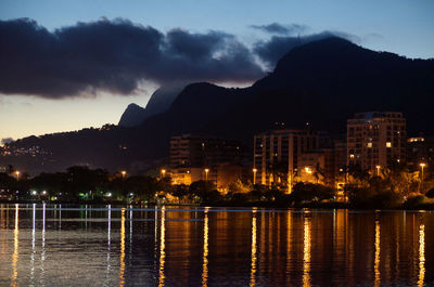 Illuminated city by lake against sky at sunset