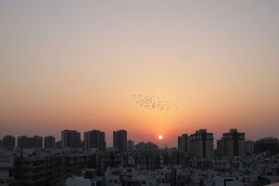 Birds flying over buildings against sky during sunset