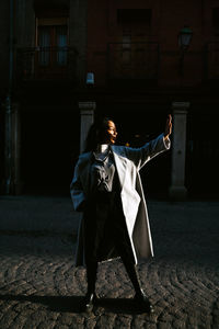 Full body of young ethnic female in stylish autumn outfit outstretching hand and covering face from sunlight while standing on pavement of old urban street