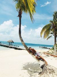 Boy lying on coconut palm tree trunk at beach against sky