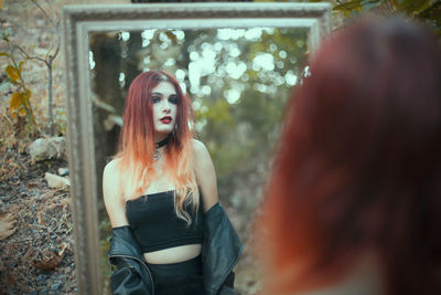 Fashionable young woman looking at her reflection in mirror at forest