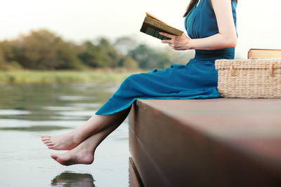 Low section of mature woman reading book sitting on pier over lake