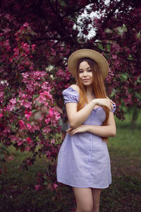 Portrait young beautiful girl teenager in a purple dress and in hat stands blooming pink apple tree