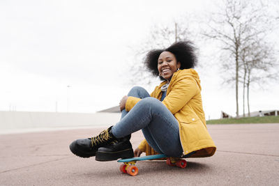 Smiling woman sitting on skateboard on footpath against clear sky