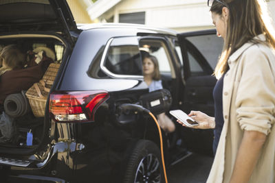 Portrait of mother, father and two daughters standing by car at electric vehicle charging station