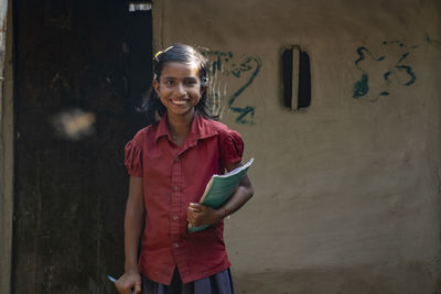 Portrait of a smiling girl student standing against wall holding book
