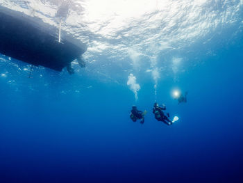 Group of professional divers in wetsuits taking picture of each other while swimming near water surface with boat undersea in deep sea