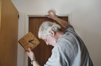 Close-up of senior man holding wall clock while standing by door on sunny day