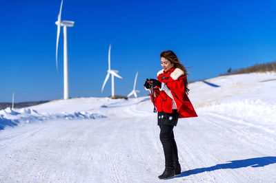 Woman holding camera while standing on snow covered road against wind turbines and clear blue sky