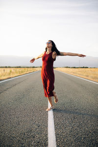 Carefree female traveler in summer dress walking along empty roadway with outstretched arms and looking away while enjoying freedom