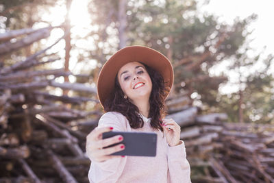 Smiling woman using smart phone while standing against woodpile in forest