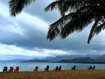 Empty adirondack chairs at beach against cloudy sky