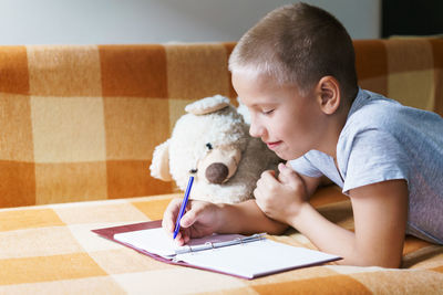 Caucasian boy lying on couch does his homework, writes with a pen in a notebook