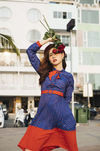 Portrait of young woman holding rose standing against buildings