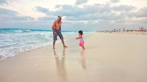 Full length of grandfather and granddaughter on beach against sky