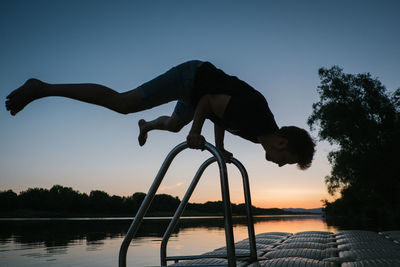 Silhouette man holding railing on pier over lake against sky during sunset