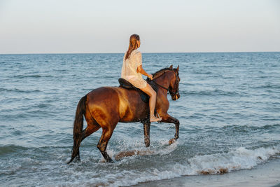 Beautiful woman in white dress riding a horse along the seashore at sunset