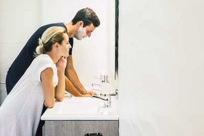 Side view of woman standing by man shaving in bathroom