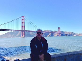 Senior man sitting on retaining wall with golden gate bridge in background against clear sky