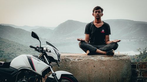 Young man sitting on mountain against sky