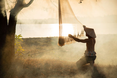 Man adjusting fishing net while sitting by tree