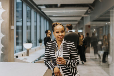 Portrait of smiling mature businesswoman wearing striped jacket holding champagne flute leaning on table at office