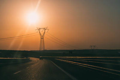 Electricity pylons by road against sky during sunset