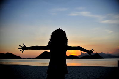 Silhouette girl with arms outstretched standing at beach against sky during sunset