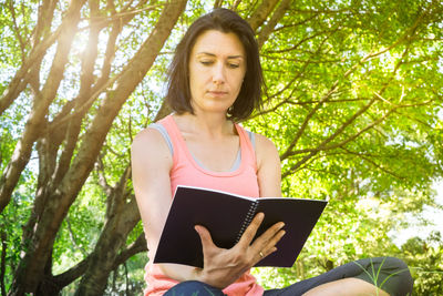 Low angle view of young woman reading book against trees