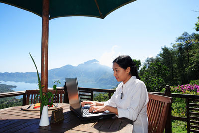 Woman using laptop while sitting against mountain and sky