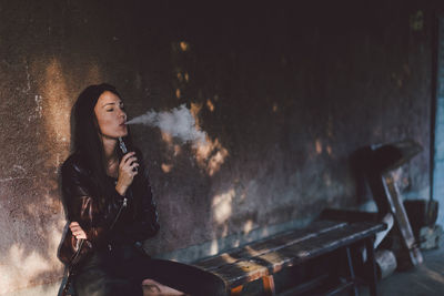 Young woman smoking electronic cigarette while sitting on bench against wall on footpath