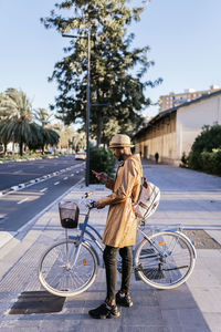 Full body side view of african american male standing on bicycle near road with crosswalk on street with buildings and trees