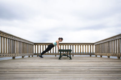 Woman doing push-ups on bench at pier against cloudy sky