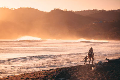 Mother and son walking on beach against sky during sunset