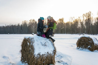 Side view of women kneeling on hay bale over snow covered land against trees during winter