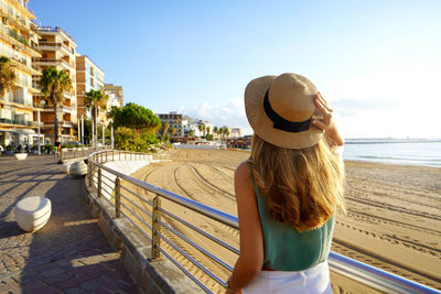 Back view of girl holding straw hat walking on crotone promenade on calabria coast, italy.