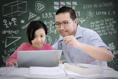 Man and girl using laptop at desk against blackboard