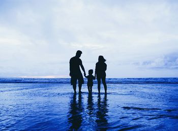 Silhouette family standing on shore at beach against cloudy sky